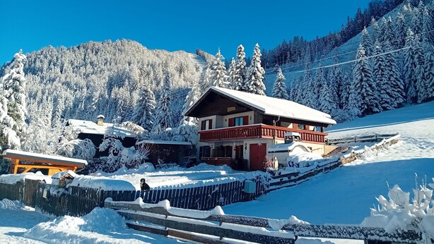 A chalet-style building with a snow-covered roof and a balcony, surrounded by a snowy landscape and a forested mountain.