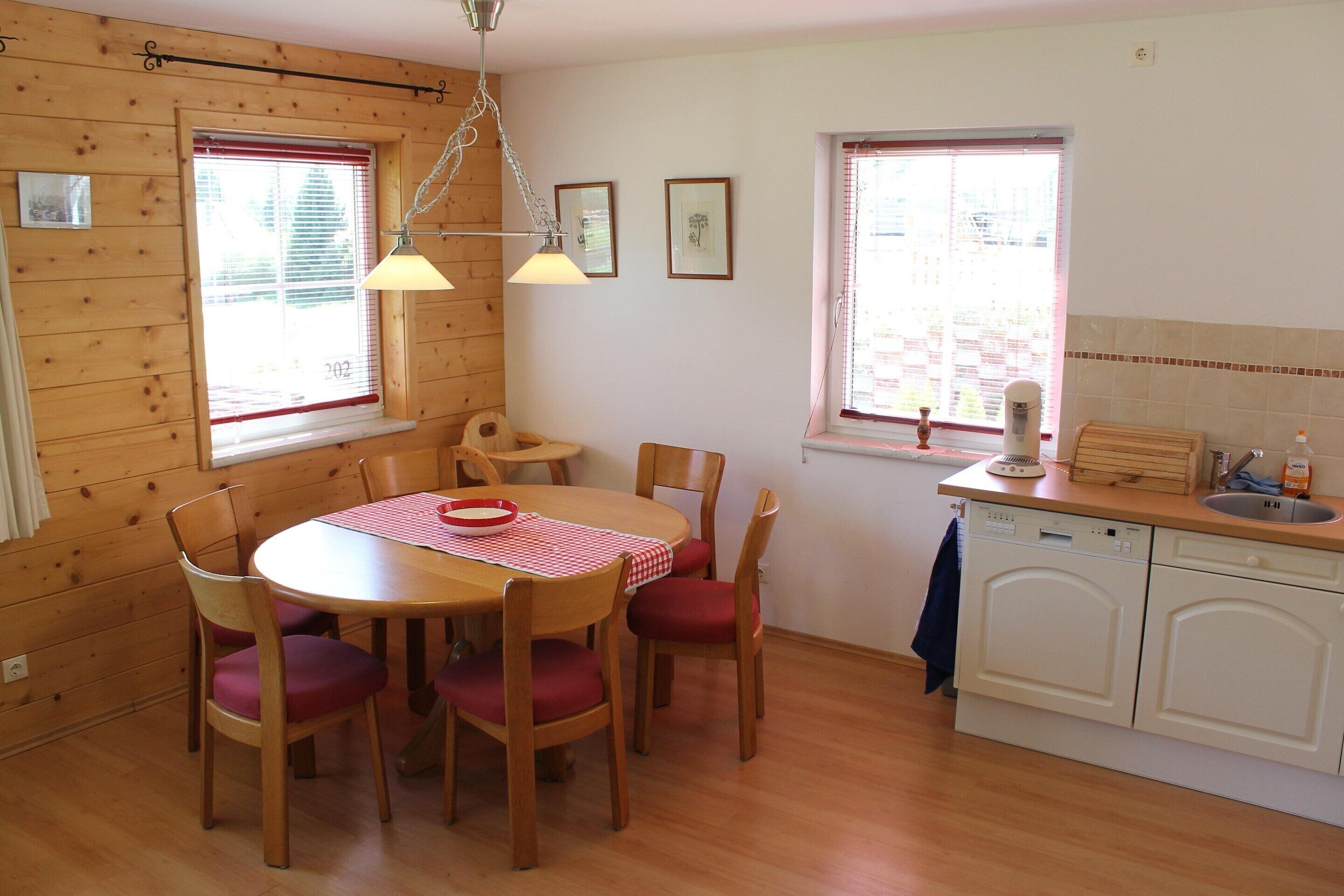 A wooden-paneled room with a dining table, chairs, a kitchen area with a sink, and two windows with blinds.
