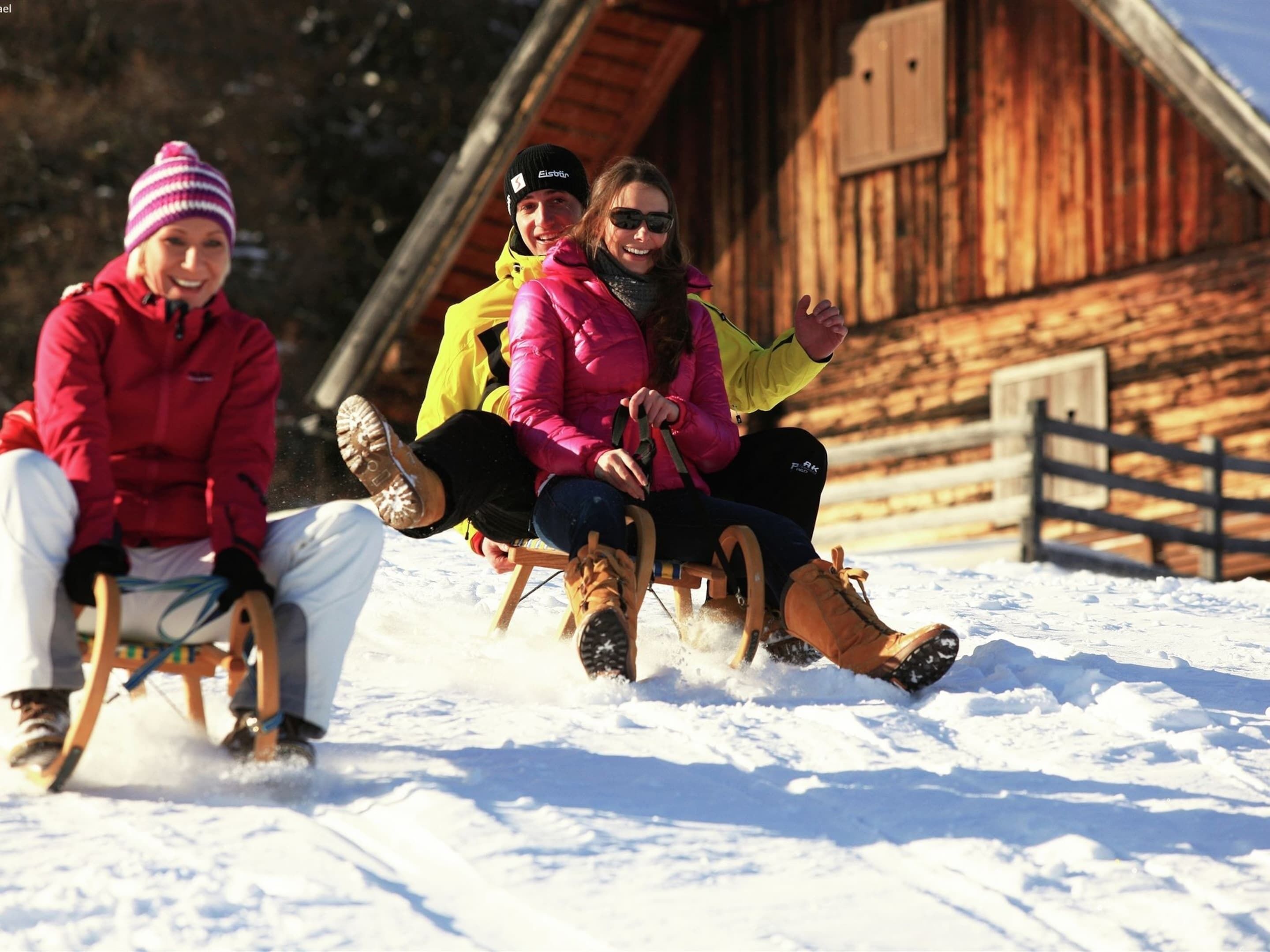 Tres personas bajan en un trineo de madera sobre nieve, con una cabaña de madera al fondo.