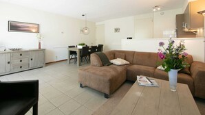 A modern living room with a sectional sofa, a dining table with chairs, a kitchen area, and a small cabinet. - Relaxing Woodland Retreat (Rheezerveen)