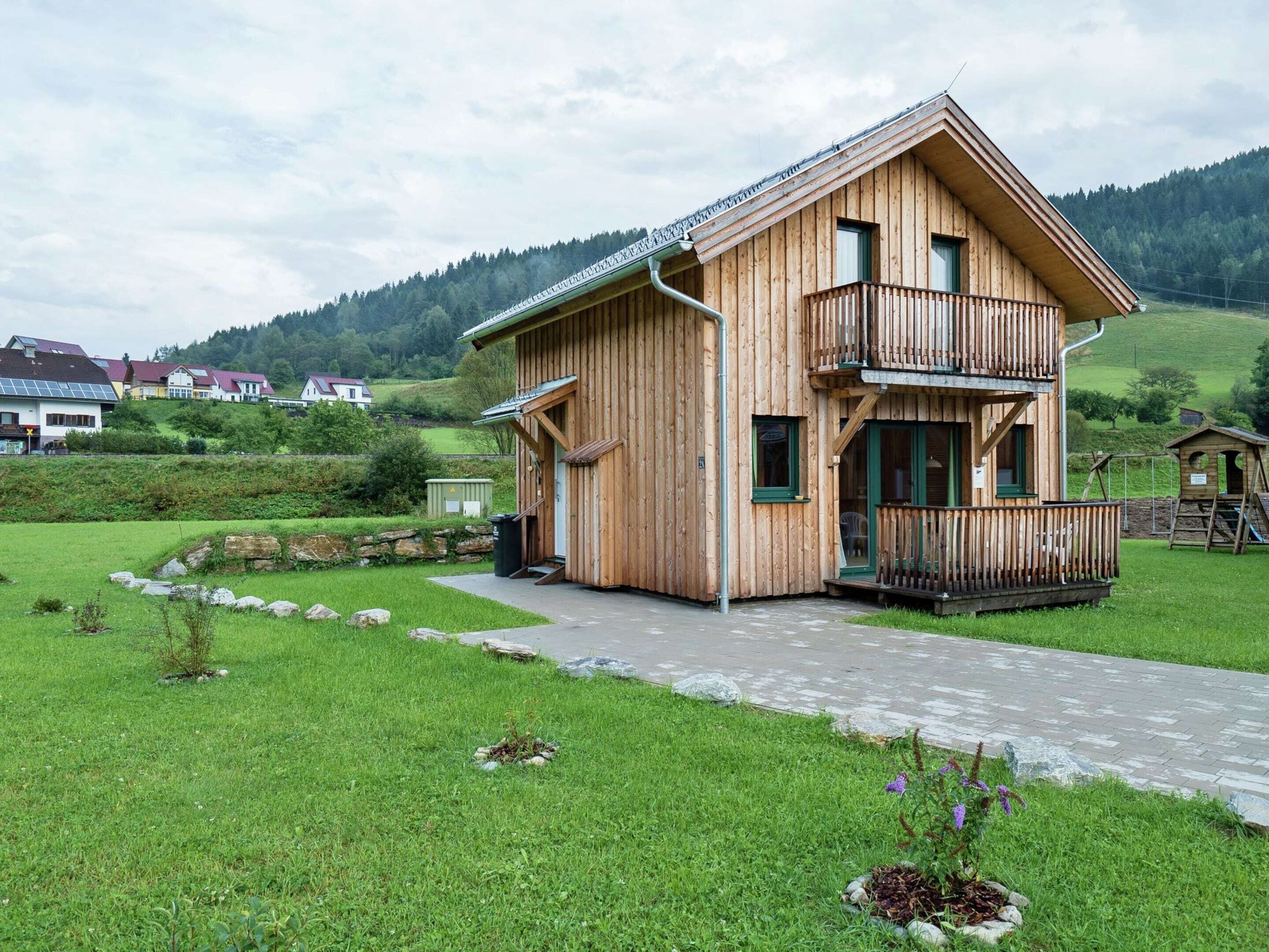 Een houten chalet met balkon, gelegen in een groen landschap met een stenen pad dat naar de ingang leidt.