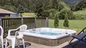 A hot tub on a wooden deck with two white chairs, surrounded by a wooden fence, overlooking a mountainous landscape.