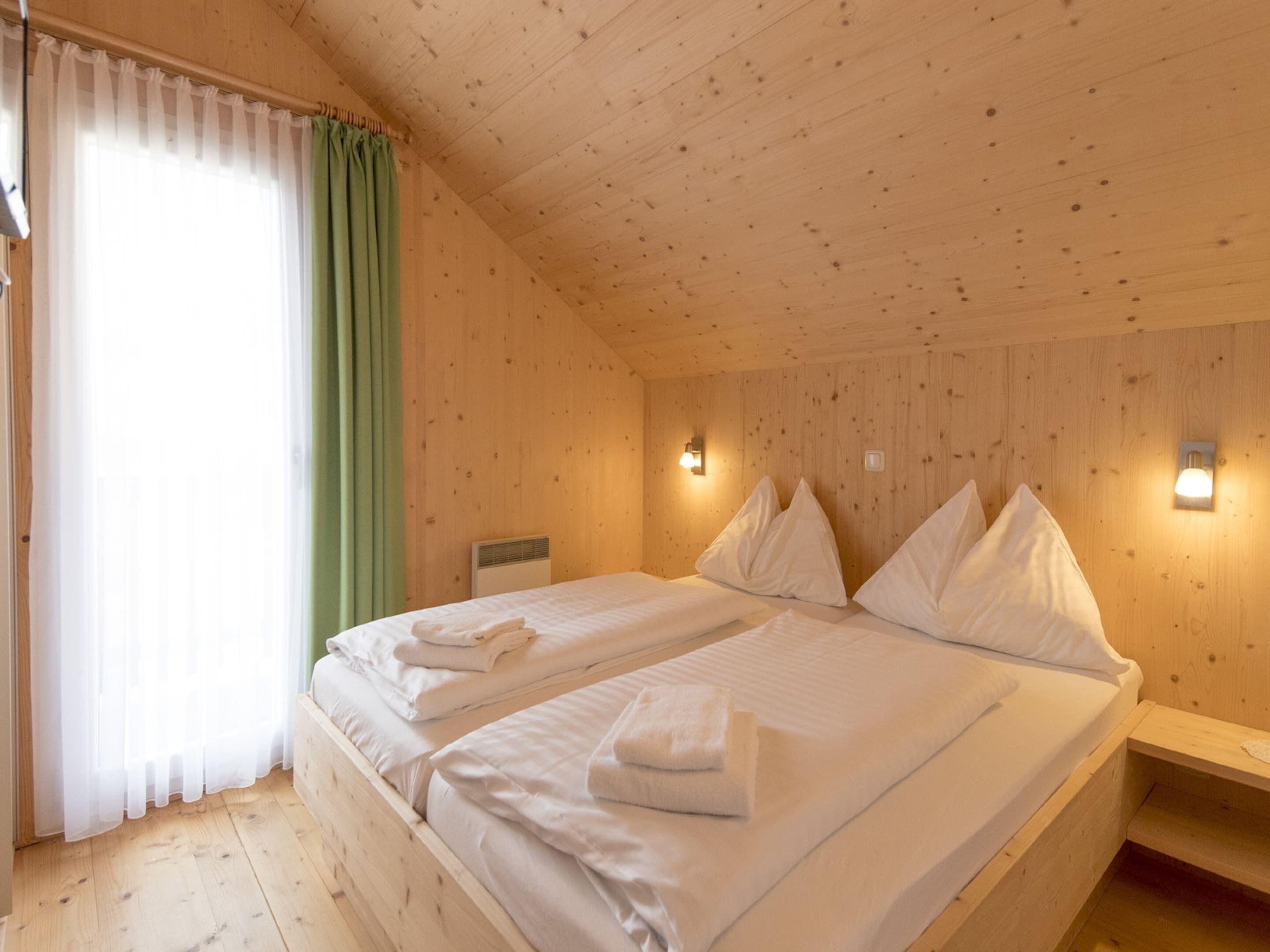 A wooden cabin bedroom with two beds, a window with green curtains, and wall-mounted lights.