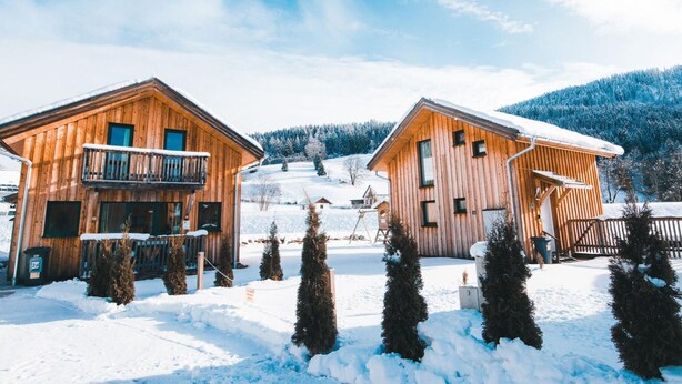 Two wooden cabins with balconies, surrounded by snow-covered ground and trees, set against a mountainous backdrop.