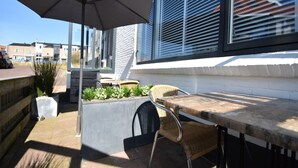 Eine Terrasse im Freien mit Tisch und Stühlen, einem Betonpflanzgefäß mit Pflanzen und einem großen Sonnenschirm.