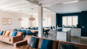 Un vaste espace de vie comprenant un grand canapé marron, un coin repas avec plusieurs tables et chaises, et une cuisine dotée d’un grand îlot central.