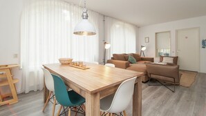 A modern living room with a wooden dining table, white and teal chairs, a brown sofa, and a large window with sheer curtains.