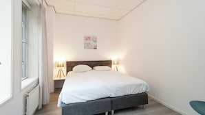 A bedroom with a bed, bedside lamps, a window with curtains, a radiator, and a wall-mounted artwork.