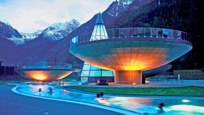A modern architectural structure with a large, circular, glass-bottomed pool in the foreground, surrounded by a paved walkway.