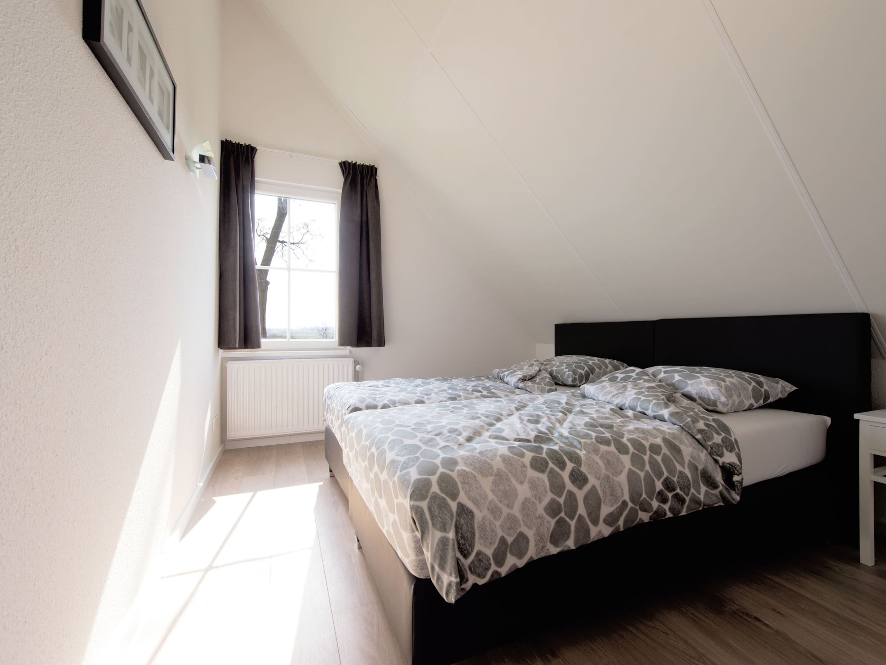 A bedroom with a bed, a window with curtains, a radiator, and a wall-mounted picture.