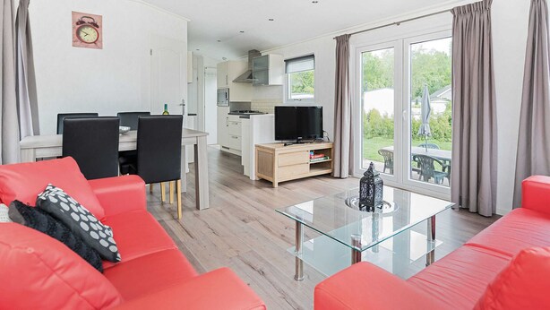 A modern living room with red sofas, a glass coffee table, and a kitchen in the background. - Chalet in Parc de Kievit With Private Garden (Baarle-Nassau)
