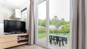 A modern living room with a flat-screen TV on a wooden console, a sliding glass door leading to an outdoor patio with a table and chairs. - Chalet in Parc de Kievit With Private Garden (Baarle-Nassau)