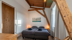 Une chambre avec un plafond en bois, un lit avec une literie grise, une commode en bois et une chaise en osier.