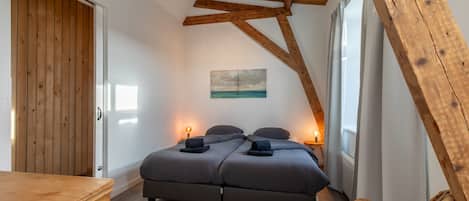 A bedroom with a wooden ceiling, a bed with grey bedding, a wooden dresser, and a wicker chair.