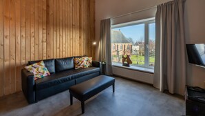 Une pièce avec un mur en bois, un canapé en cuir noir agrémenté de coussins colorés, un banc, une grande fenêtre avec des rideaux et un téléviseur posé sur un meuble.