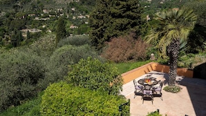 Un patio con una mesa y sillas, rodeado de exuberante vegetación y vistas a un pueblo en la ladera.