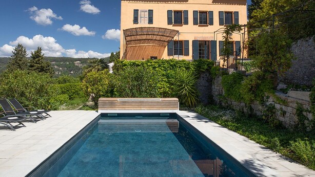 Una piscina con sillones de descanso, un edificio grande con techo de tejas y abundante vegetación.