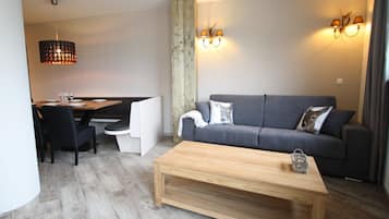A modern living room with a grey sofa, a wooden coffee table, a dining area with a wooden table and chairs, and a wall-mounted light fixture.