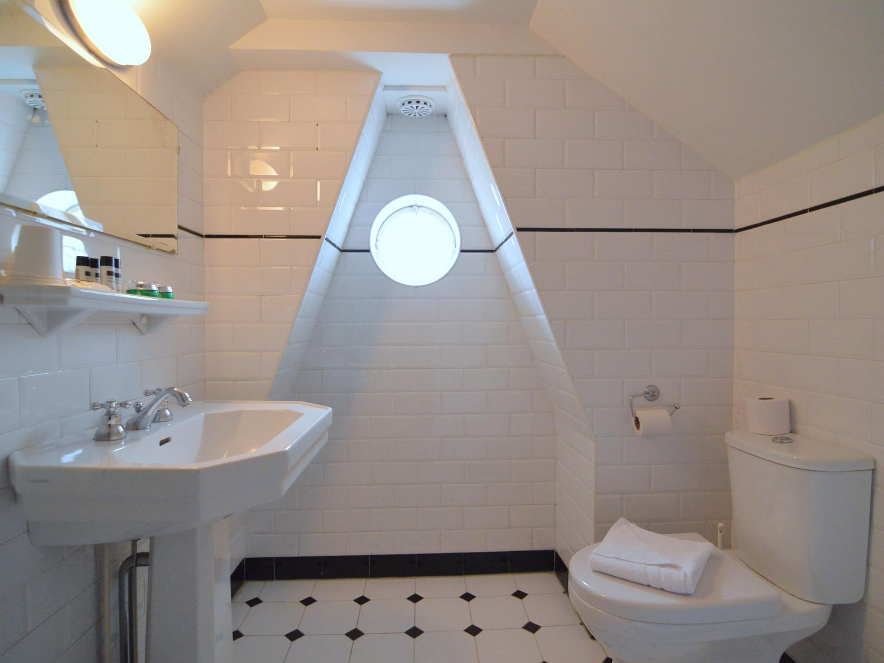 Een badkamer met een witte wasbak, toilet en spiegel. Er is een plank met toiletartikelen en een handdoek op het toilet. De vloer heeft een zwart-wit tegelpatroon.