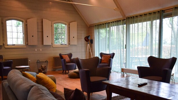 A cozy living room with a sofa, armchairs, a coffee table, and a floor lamp. There are windows with curtains, a radiator, and a wooden floor. - Charming Holiday Home in Hulshorst With Pool (Hulshorst)