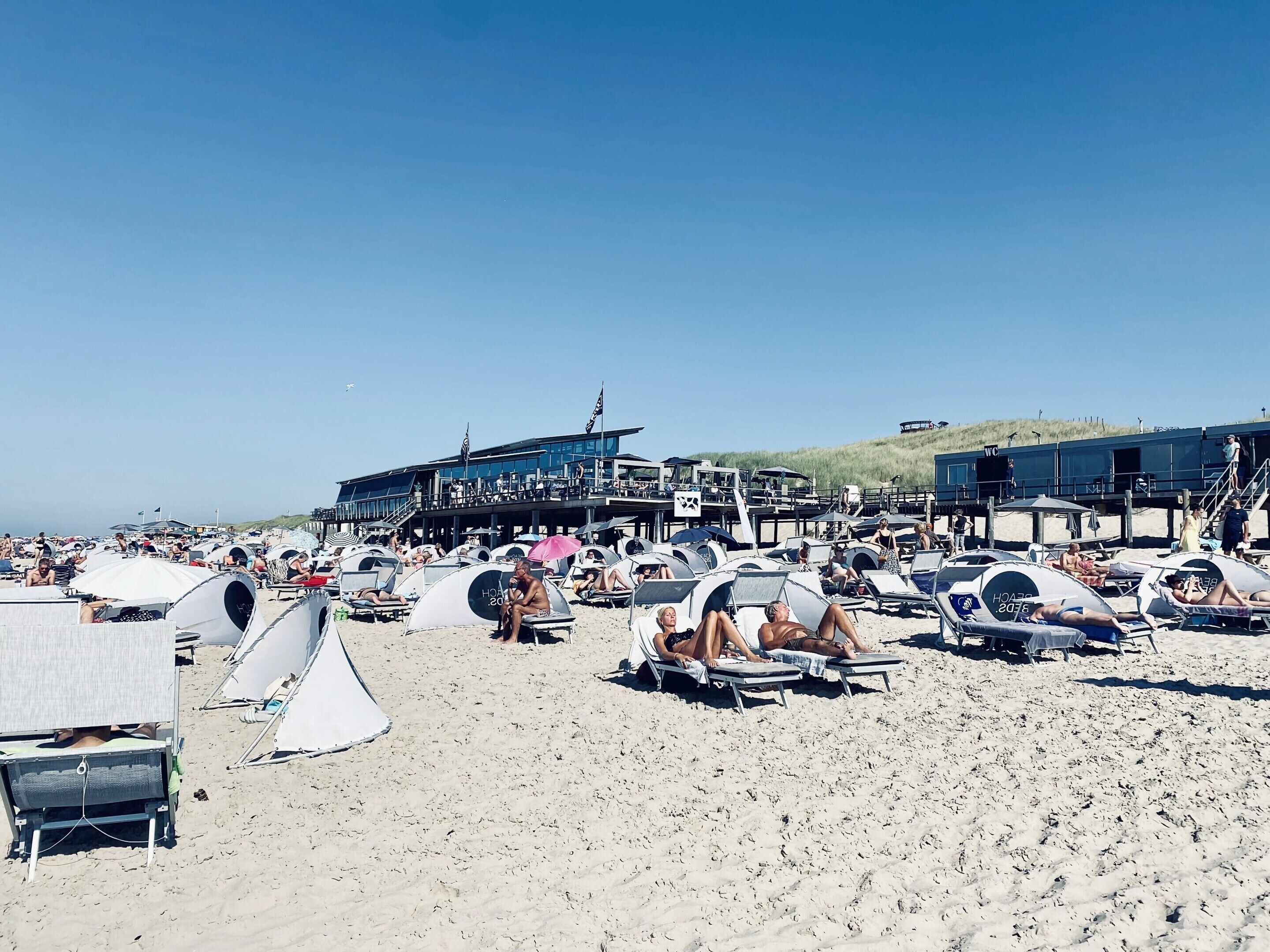 Ein Strand mit vielen Liegestühlen und Sonnenschirmen, ein großes Gebäude mit einer Terrasse und einem klaren Himmel.