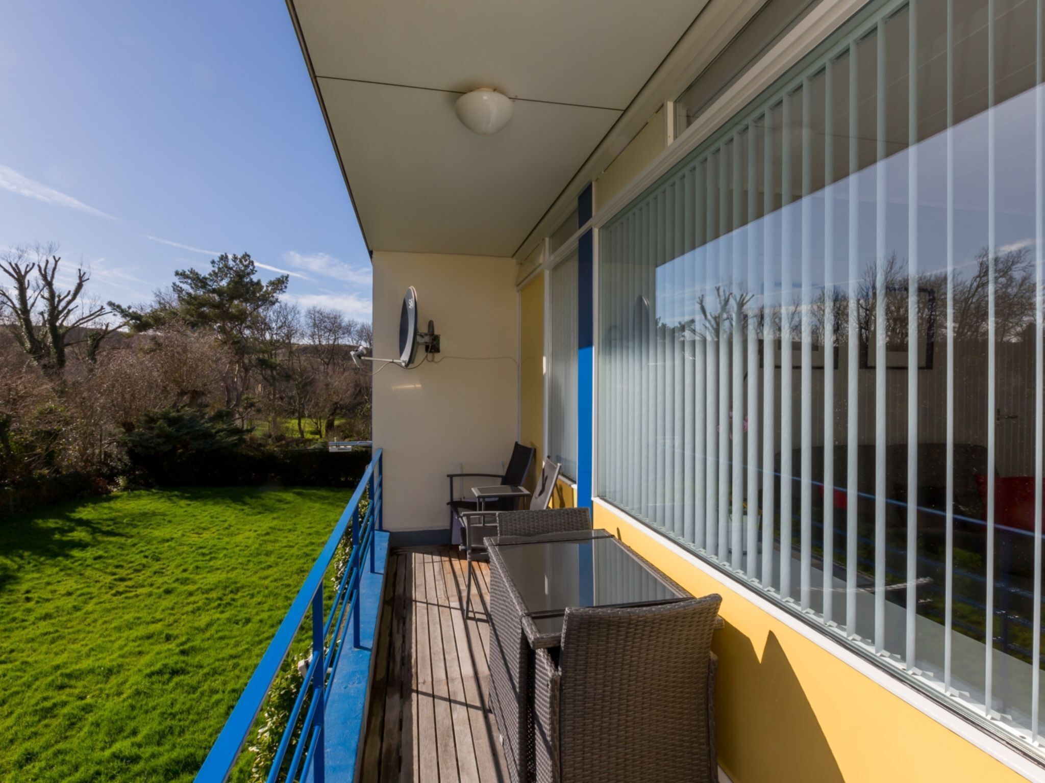 A balcony with a glass table and chairs, a blue railing, and a view of the garden and trees.