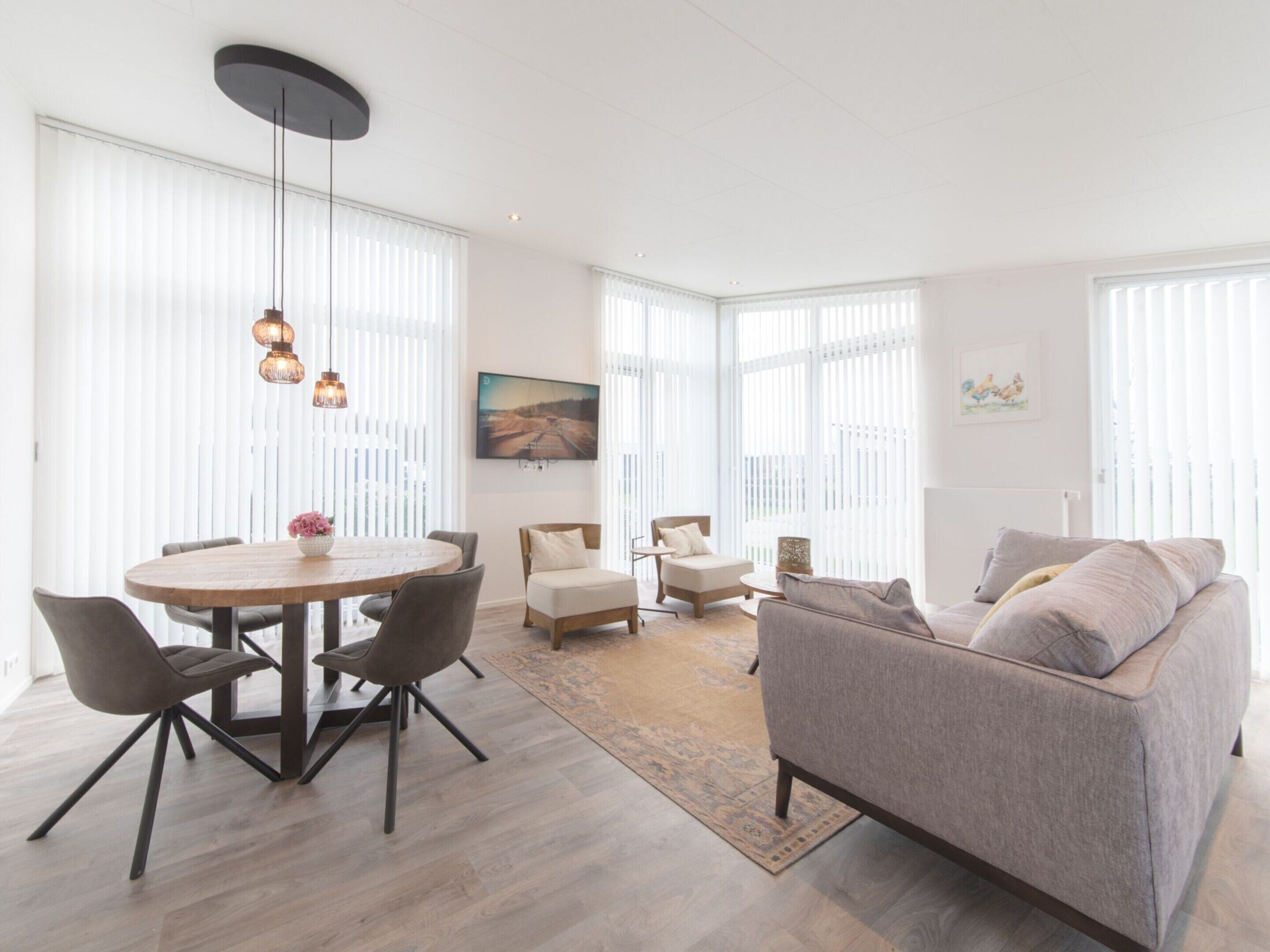 Een ruime woonkamer met een ronde eettafel, moderne stoelen, een grijze bank en grote ramen met zonwering.
