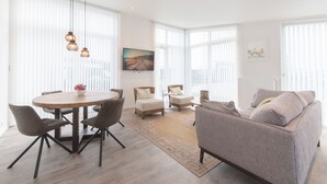 A spacious living area with a round dining table, modern chairs, a grey sofa, and large windows with blinds.