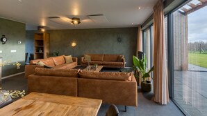 Un salon moderne avec un canapé d’angle, une table basse et une table à manger. Une grande fenêtre offre une vue sur l’extérieur.
