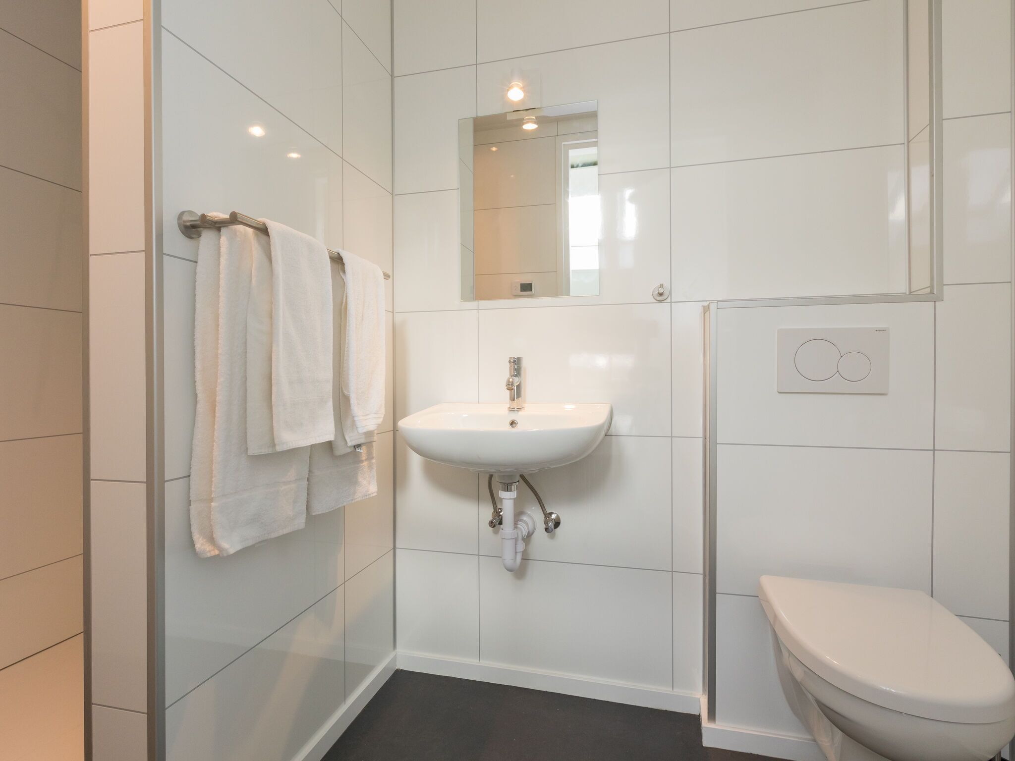 A modern bathroom with white tiles, a white sink, a mirror, and a toilet.