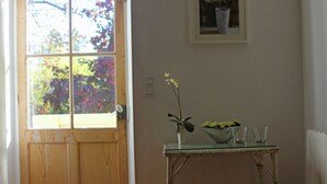 Una habitación con puerta de madera, ventana con vistas a árboles, una mesa pequeña con un jarrón y un cuenco de flores, y un cuadro en la pared.