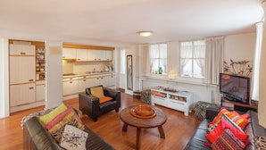 A modern living room with a sofa, a coffee table, and a television. There is a kitchen area with cabinets and a refrigerator. - Holiday Home in Soerendonk With Garden (Soerendonk)