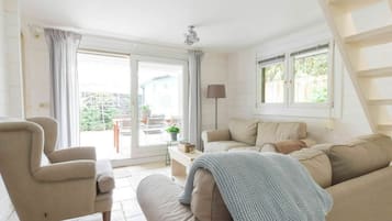 Een woonkamer met een bank, fauteuil en een tafel met een lamp. Er is een schuifdeur die leidt naar een buitenruimte met een terras en uitzicht op groen.