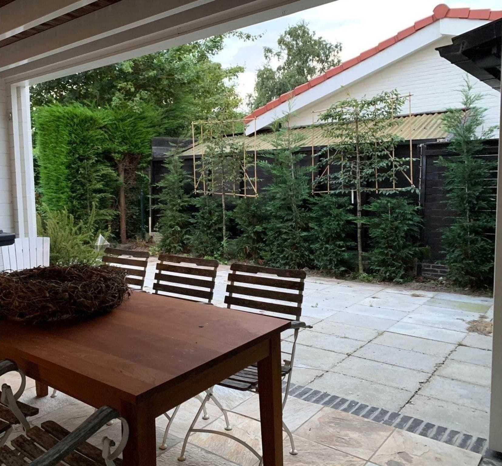 Een overdekt terras met een houten tafel en stoelen, omringd door weelderige begroeiing en een gebouw met een rood dak.