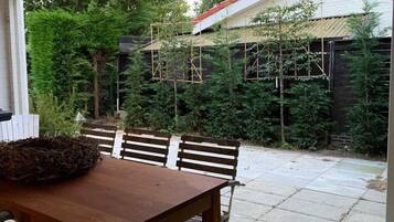 Een overdekt terras met een houten tafel en stoelen, omringd door weelderige begroeiing en een gebouw met een rood dak.