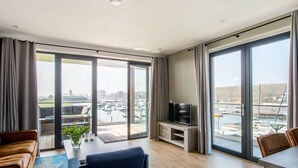 A modern living room with a large TV, a dining area, and a view of a marina through large windows. - Holiday Home in Kamperland (Kamperland)