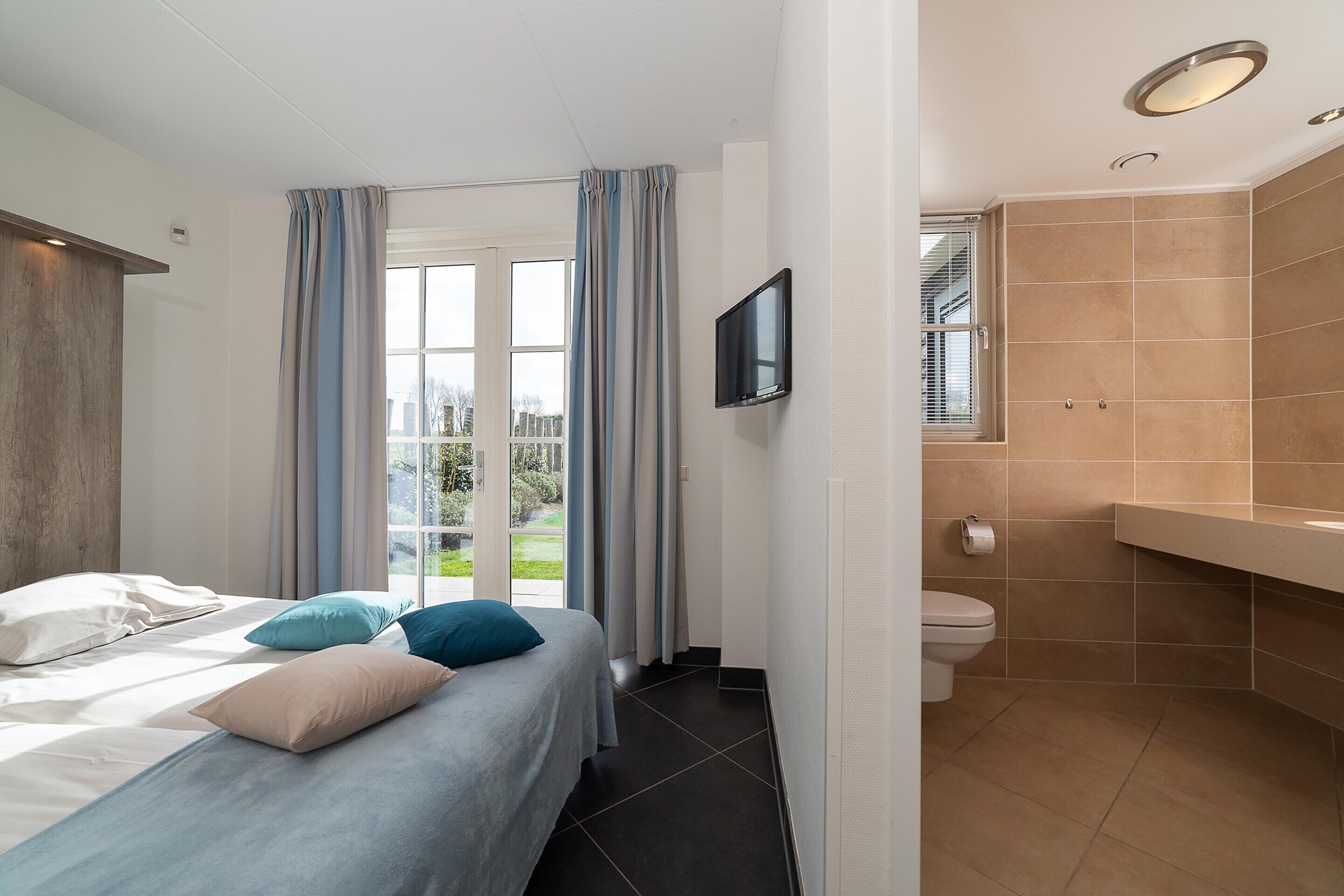 A hotel room with a bed, a television, a bathroom with a toilet and sink, and a view of the outdoors.