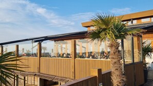 Eine Holzterrasse mit Glasgeländern, die auf einen Strand, Palmen und ein Gebäude mit der Aufschrift 'REY BLAN' blickt.