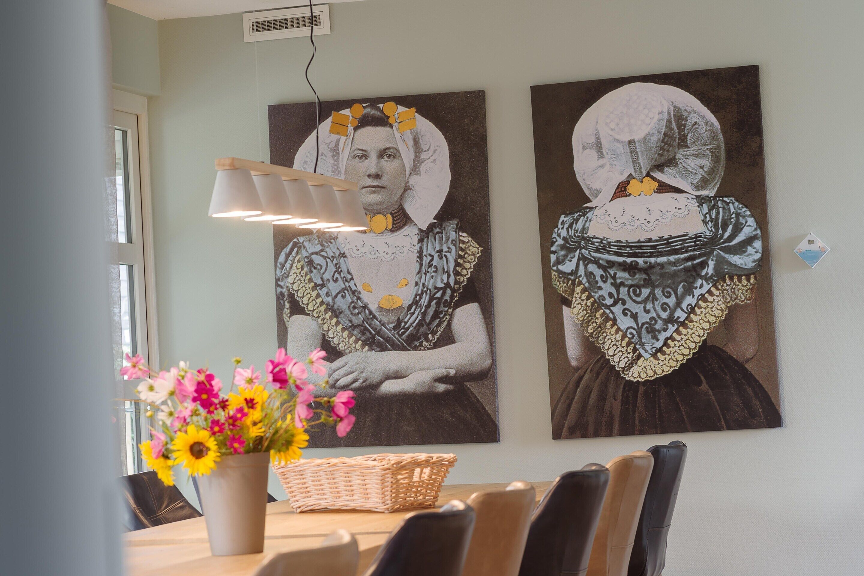 A dining area with a table set for four, a vase of flowers, and two wall-mounted paintings of women in traditional attire.