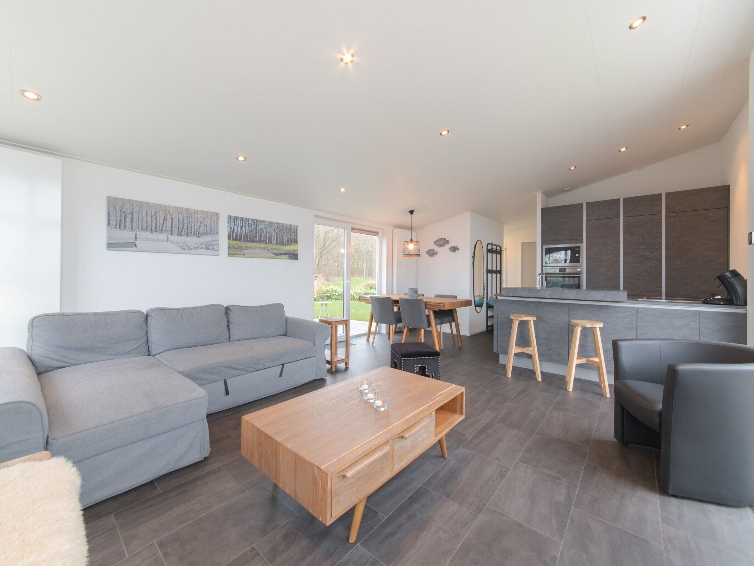 Ein modernes Wohnzimmer mit grauem Sofa, Couchtisch aus Holz und einem Essbereich mit Stühlen und einem kleinen Tisch.