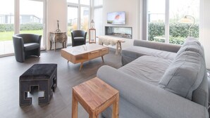 A spacious living room with a grey sofa, a wooden coffee table, and two armchairs. A television is mounted on the wall, and there is a large window providing a view of the outdoors. - Modern Chalet Near Beach (Wemeldinge)
