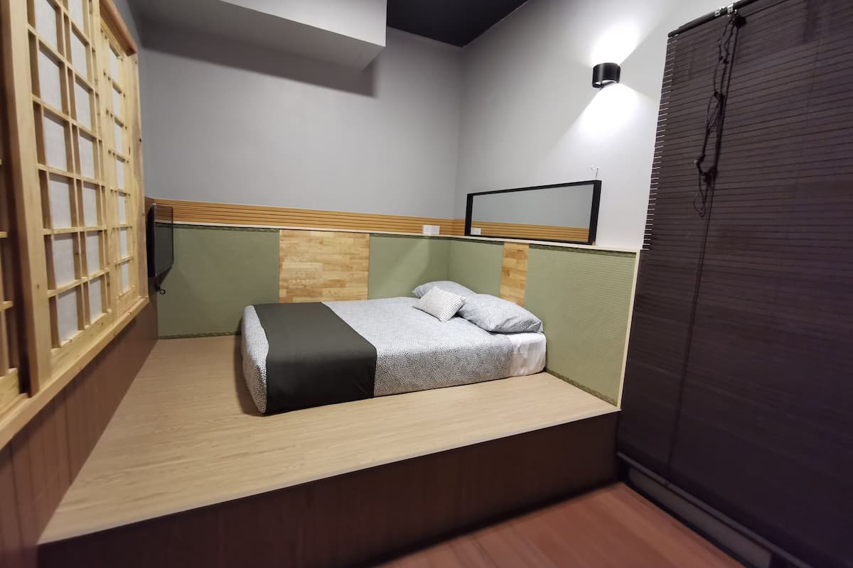 Een kleine kamer met een bed, een houten hoofdbord, een wandlamp en een raam met zonwering.