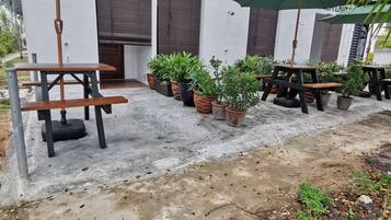 Un área de estar al aire libre con mesas y sillas, rodeada de plantas en macetas y sombrillas.