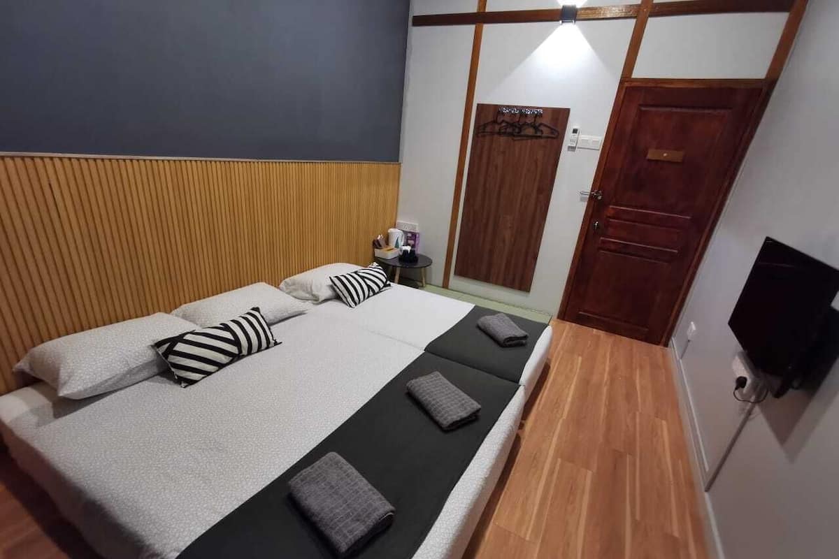 Een keurig opgemaakt bed met kussens en een deken, een nachtkastje met een lamp, een houten hoofdbord, een houten deur en een aan de muur bevestigde televisie.