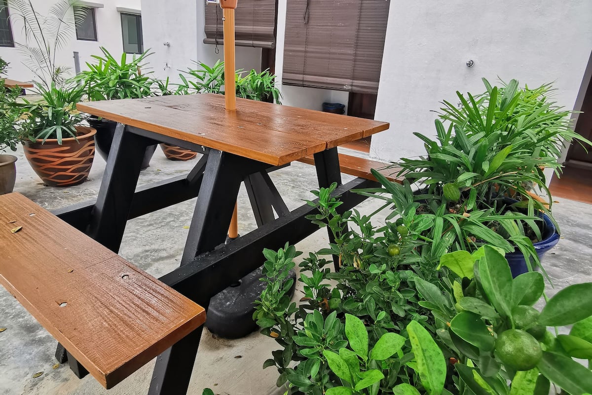 Een houten tafel en bankje onder een groene parasol, omringd door potplanten.