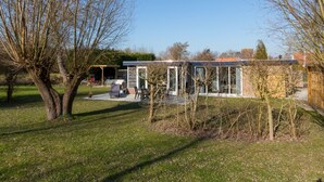 A modern house with a glass conservatory, a patio with outdoor furniture, and a garden with trees. - Holiday Home Near Veerse Meer Lake Wolphaartsdijk (Wolphaartsdijk)