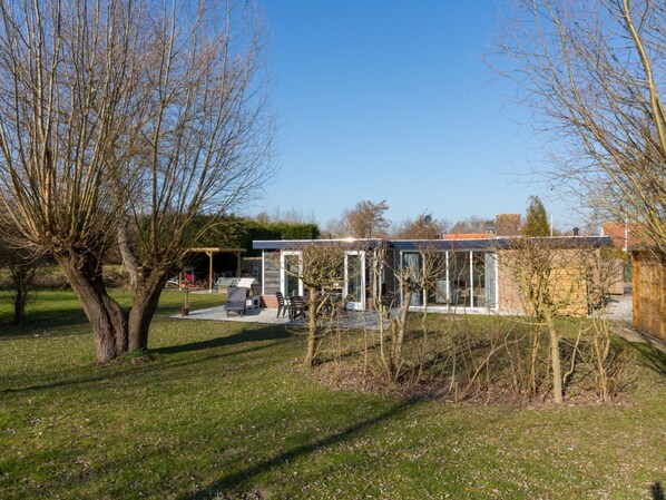 A modern house with a glass conservatory, a patio with outdoor furniture, and a garden with trees. - Holiday Home Near Veerse Meer Lake Wolphaartsdijk (Wolphaartsdijk)