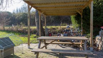 A wooden picnic shelter with benches, surrounded by a garden and a bicycle rack.