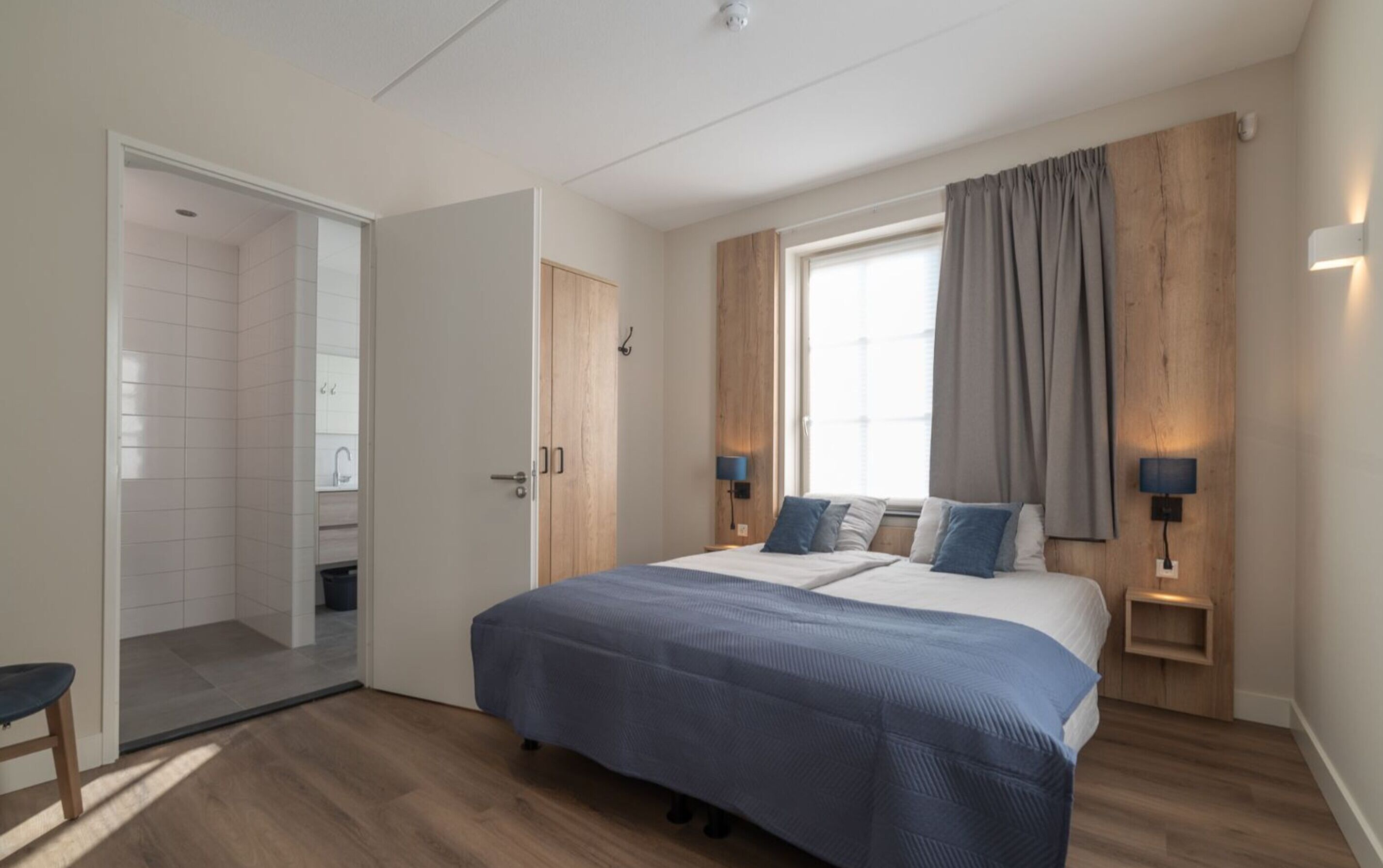 A modern bedroom with a large bed, wooden headboard, two bedside lamps, and a window with curtains.