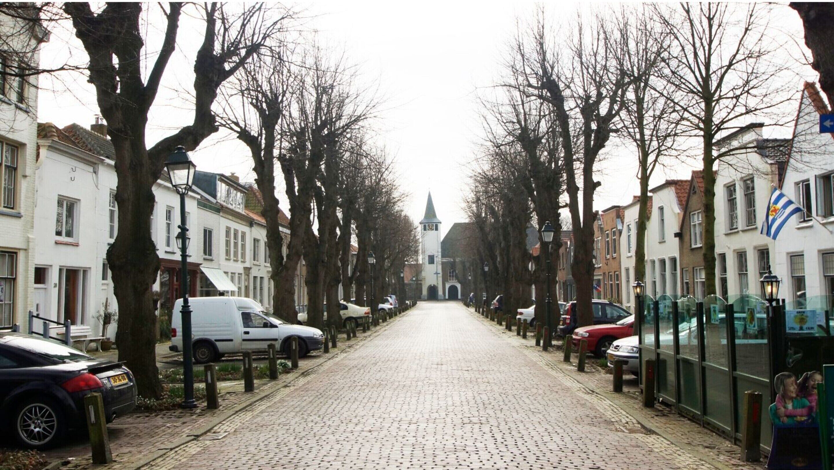A cobblestone street lined with parked cars and leafless trees, leading to a church with a tall steeple.